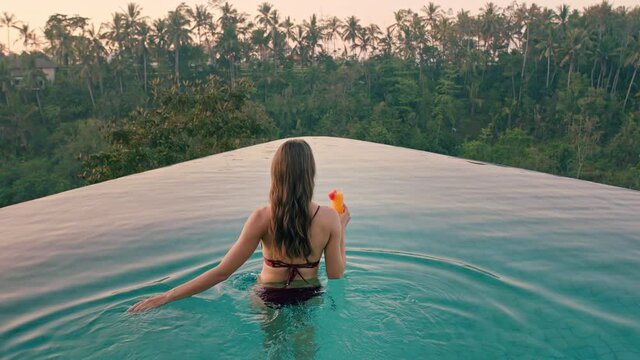 beautiful woman in swimming pool drinking cocktail enjoying luxury lifestyle on exotic summer vacation at hotel spa with view of tropical jungle at sunrise