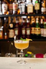 Glass with yellow lemon cocktail decorated with dried pineapple and mint on the blurred bar counter background.