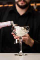 male bartender pouring to the measuring glass cup with ice cream an alcoholic drink from steel jigger on the bar counter on the blurred background