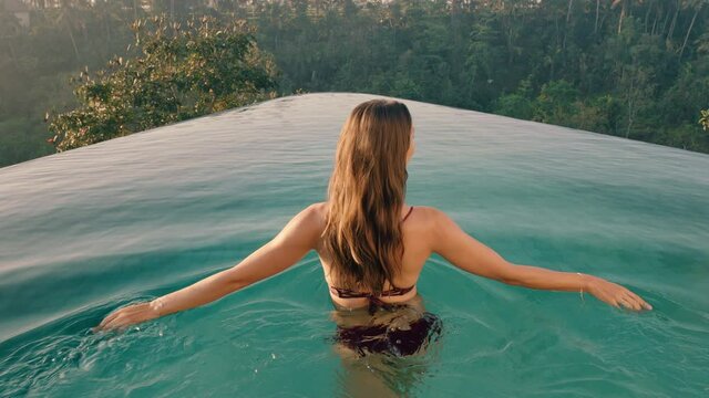 sexy woman walking in swimming pool enjoying luxury lifestyle on exotic summer vacation at hotel spa with view of tropical jungle at sunrise