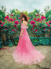 Obraz na płótnie Canvas fantasy woman in pink vintage style boho dress, stands. Fashion model girl back rear view. Straw hat wide brim. Summer nature green grass trees bushes blooming rose flowers. elegant long evening gown.