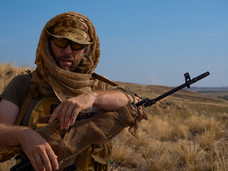 Close-up photo of a mercenary sniper in camouflage clothes under the scorching sun. He stands with...