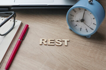 Work, Life Balance concept. The word REST on the wooden table with an alarm clock, pen, and notebook at the side