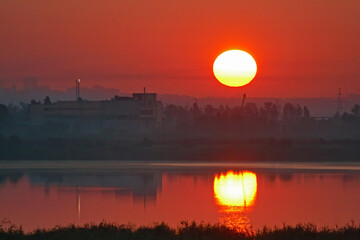 View of industrial buildings and structures in predawn fog and red sky with large sun disk