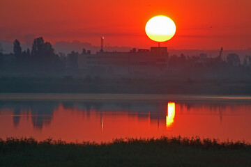 View of industrial buildings and structures in predawn fog and red sky with large sun disk