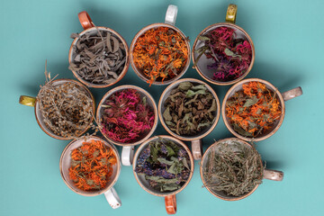Fototapeta premium Assortment of dried relaxing tea herbs in colourful cups on blue background close up. Calendula, mint, anise hyssop, monarda didyma, wormwood, sage leaves.