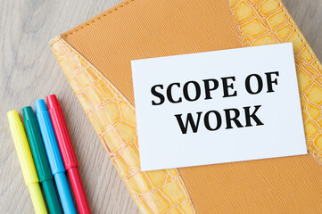 a card with text SCOPE OF WORK on a yellow notebook on the table next to colored markers