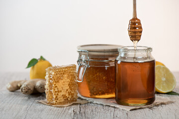 Honey in a jars, piece of bee comb and honey dipper on light background. Next to it is sliced lemons and ginger. Home treatment for flu. Honey harvesting concept.