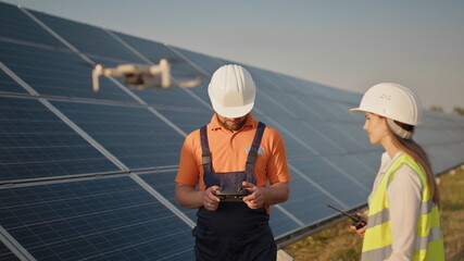 Industrial expert wearing helmet and controlling drone in photovoltaic solar power plant. Solar panel array installation. Technologies and ecology. Female investor checks the work