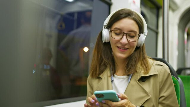 Young caucasian woman wearing headphones listening music on the smartphone and dancing while going somewhere in the tram and enjoying. Transportation and technologies concept