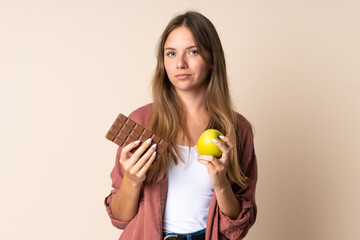 Young Lithuanian woman isolated on beige background taking a chocolate tablet in one hand and an apple in the other
