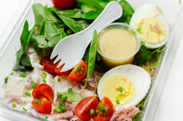 Healthy salad in the plastic food box, potato salad with hard boiled egg, shredded ham, cucumber, tomatoes, lettuce and spinach with a honey and mustard dressing pot