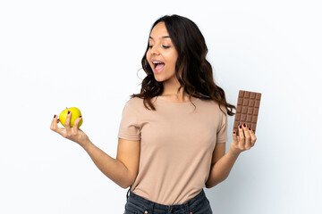 Young woman over isolated white background taking a chocolate tablet in one hand and an apple in...
