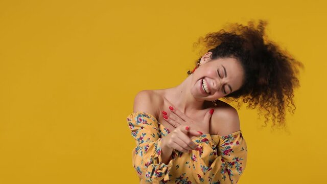Excited cheerful fun charming vivid young latin curly woman 20s wears casual flower dress look camera laugh smiling watch comedy movie pointing index finger on you isolated on plain yellow background