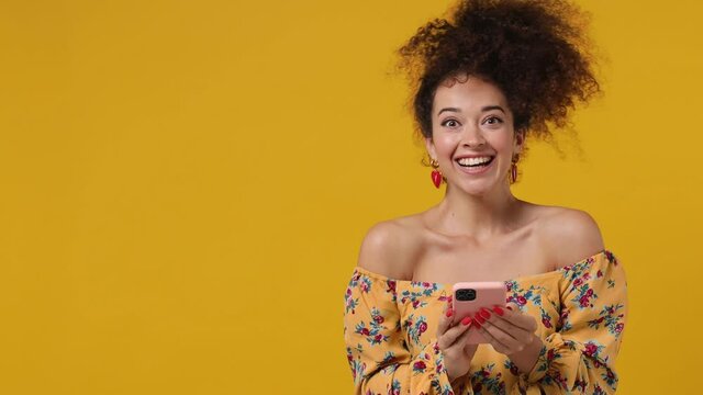 Surprised promoter young latin curly woman 20s wears casual flower dress hold use mobile cell phone look aside on workspace copy space mockup promo commercial area isolated on plain yellow background