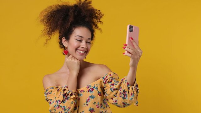 Charming vivid amazing marvelous magnificent young latin curly woman 20s wears casual flower dress doing selfie shot on mobile phone post photo on social network isolated on plain yellow background