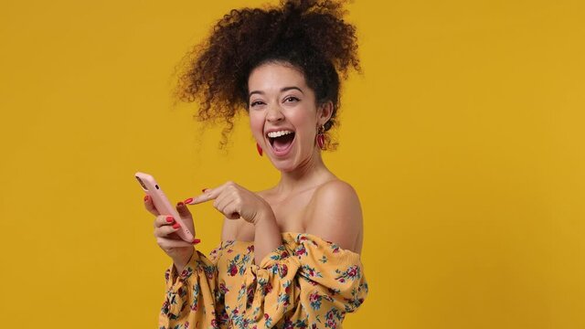 Satisfied young latin curly woman 20s wear casual flower dress hold use mobile cell phone typing browsing swipe chatting send sms doing online shopping booking tour isolated on plain yellow background