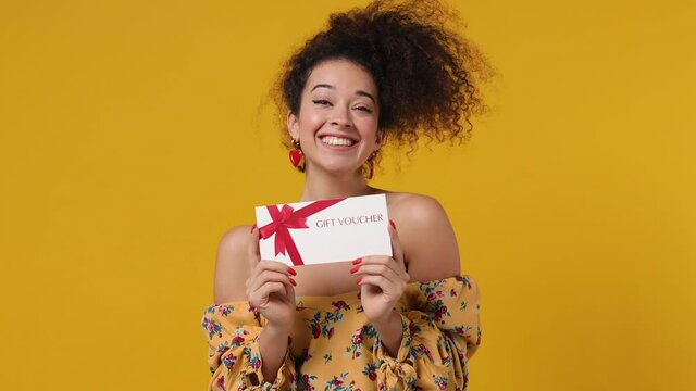 Surprised shocked happy vivid cheery young latin curly woman 20s wear casual flower dress pointing finger on gift certificate voucher for store doing winner gesture isolated on plain yellow background