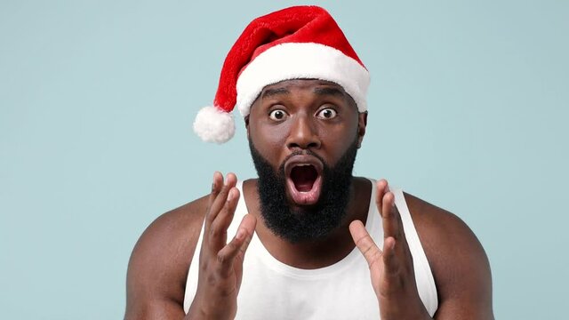 Merry surprised fun young african american Santa man in white tank top Christmas hat say what wow omg no way isolated on plain light blue background studio portrait. Happy New Year celebration concept