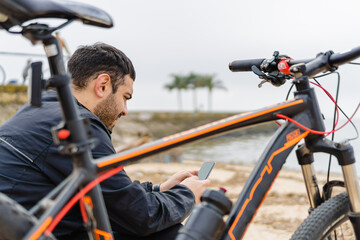 Obraz premium young latin hispanic man checking his cell phone in a park by the river next to his bicycle.