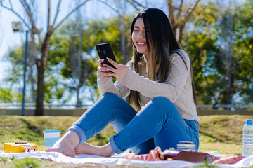 smiling, barefoot Hispanic latin young woman is chatting on top of a picnic blanket in a city park...