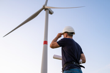 Engineer in hard hat holds a plan project for the construction of wind turbines in the field against the background of windmills. Generation of electricity. Alternative production of renewable green