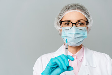 Female doctor or scientist in white medical gown, blue gloves, green cap and mask holds a syringe in hands on white background. She is ready to give an injection