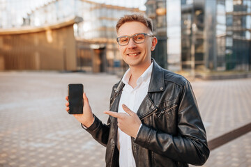 Handsome man in glasses with a smartphone on the street of a big city. Businessman points finger at phone screen on urban background