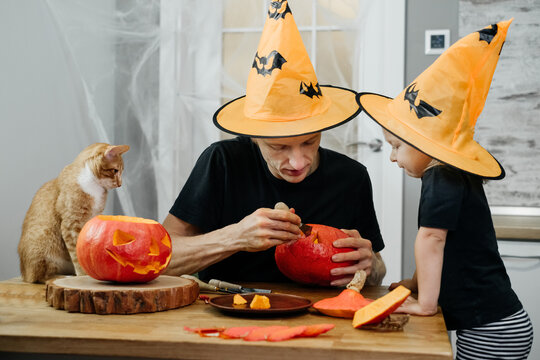 Parent with chld are carving jack o lantern from pumpkin for Halloween at home. Father in hat preparing for the selebration with daughter. 