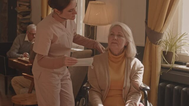 Slowmo shot of caring nurse putting protective mask on face of old grey-haired woman in wheelchair at nursing home
