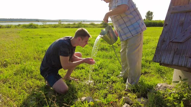 Young farmer washes his face with well water, which is poured out of bucket by an elderly man at sunset in field. High quality 4k footage