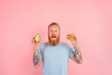 Amazed man with beard and tattoos is undecided if to eat an avocado or an hamburger