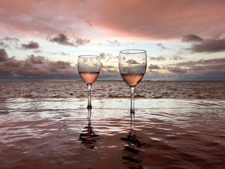 Portrait view of two half wine glasses with a sunset background in an infinite pool with oceanfront view.