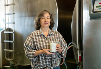 Confident elderly woman milking farm owner standing near large tanks in storage room, holding glass with fresh milk © JackF