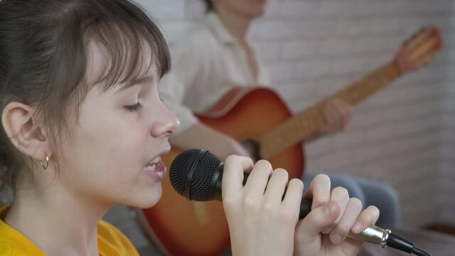 A young girl sings into a microphone with a guitar. The teenage daughter sings, the mother plays the guitar.