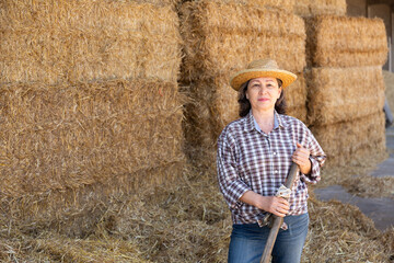Naklejka na ściany i meble Portrait of smiling adult woman farmer in straw hat posing on hayloft at cow farm