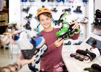 happy male shop assistant helping positive european boy to choose roller-skates in sports store.