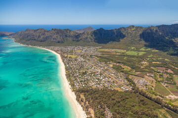 Fototapeta premium Aerial view of the coastline of the island of Oahu in Hawaii, USA. Turquoise blue water with visible reefs on left of frame, housing with green and brown mountains on the right side of the frame.