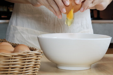 Close-up front view footage, a female cook in a white apron is cracking an egg into a cup to prepare a meal on a wooden table in the home's kitchen. Eating egg yolks is a healthy breakfast.