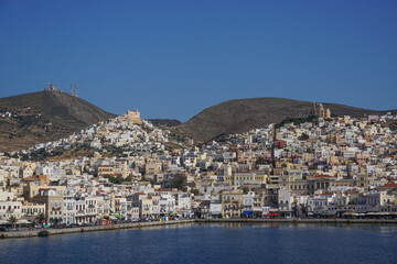 Fototapeta na wymiar Syros, Greece: The hills, houses, churches, shops, and harbor of Ano Syros and Hermoupolis shine in the sun on the Aegean island of Syros.