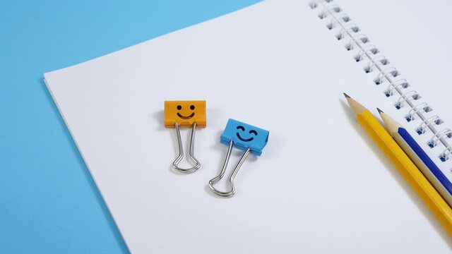 Blank paper notepad with yellow and blue binder clips on blue background. Spiral notebook and smile binder clips on table. Office supplies or education concept