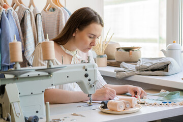 Young woman fashion designer sitting at her workplace with sewing machine in studio and working on...