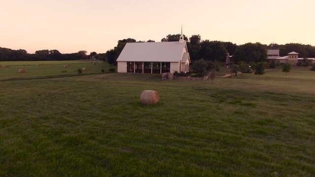 Drone footage of Small White Wedding Chapel Church on farm with bales of hay in field