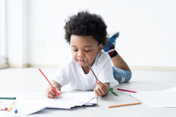 African American little boy lying and drawing on pages with colour pencils on floor at classroom. Kid learning by drawing. Education concept