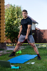 The young man goes in for sports at home in  backyard in summer day. Young sportsman with blond  hair  shakes hands with sport rubber on mat, there is  a ball, dumbeels.