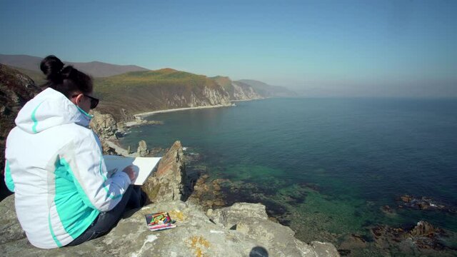 Back of a woman sitting on the cliff edge enjoying bright sunny day and paiting amazing marine landscape