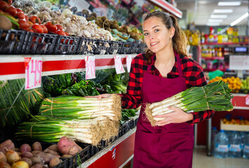 Young smiling female seller lay out leek on the counter in supermarket