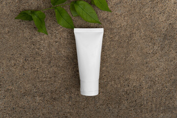 Top down view above facial skincare white tube bottle product with blank label and green leaves on sandy dark brown stone ground rough texture background. Skincare Environmental friendly concept

