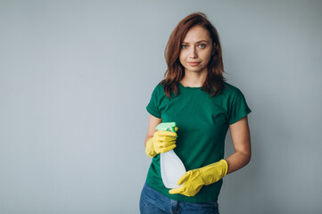 Happy smiling young woman holding spray detergent while wearing yellow rubber gloves for hands protection during cleaning isolated over grey background copyspace 