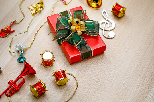 Small red paper wrapped gift present box with green and gold shiny ribbon bow tie placed on wooden table with small decorative music instruments on Christmas eve birthday night or new year festival